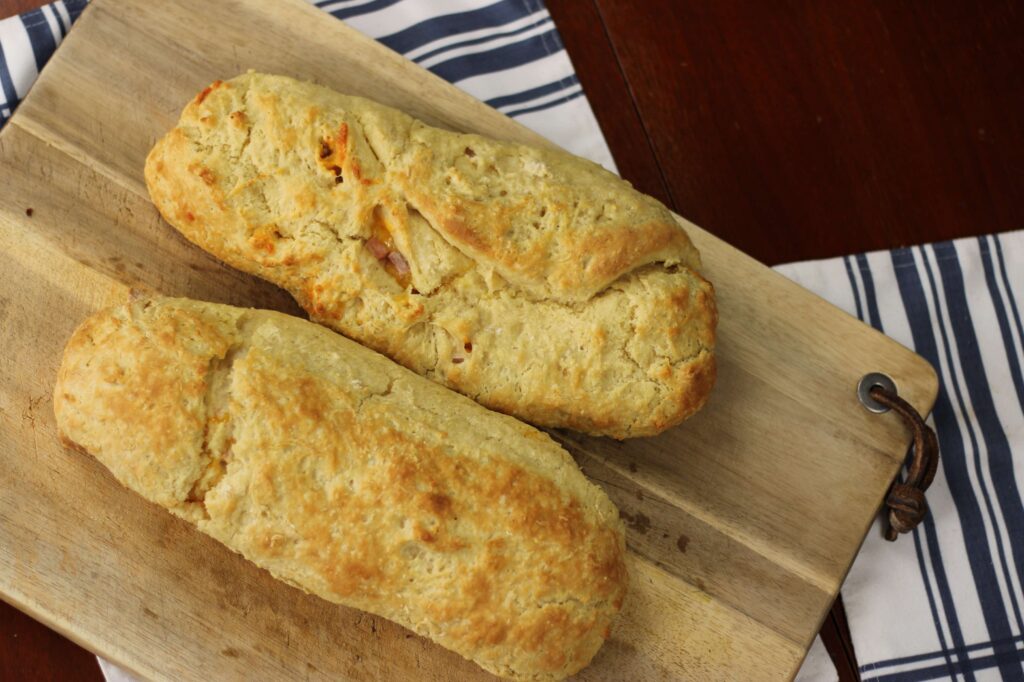 Two freshly baked ham and cheese rolls on wooden cutting board sitting on table with white napkins with navy stripes.