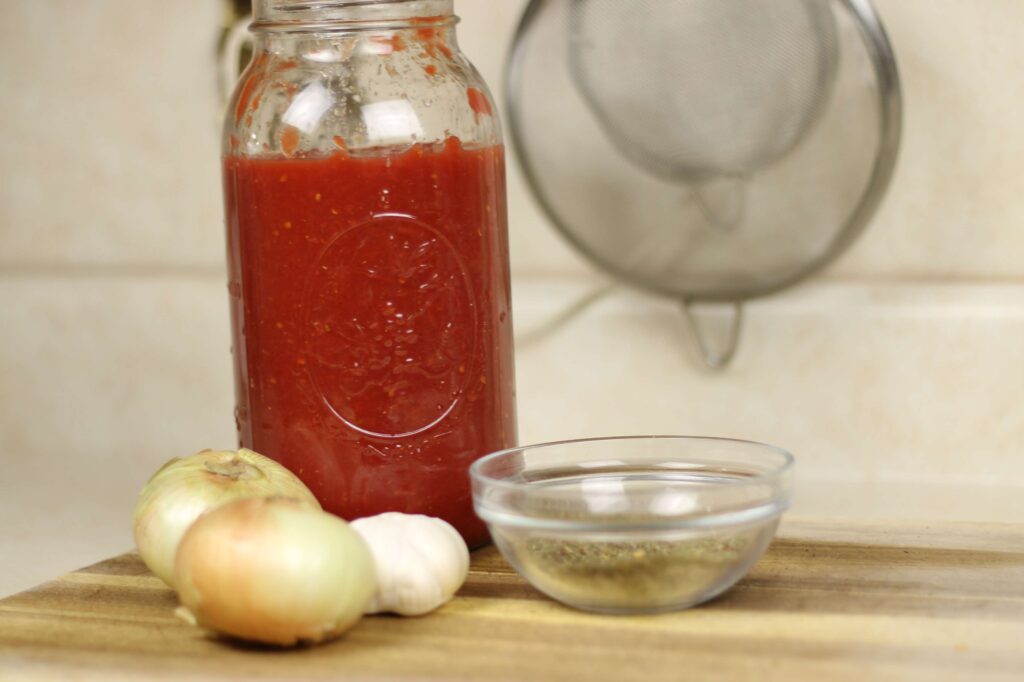 half gallon mason jar of tomato sauce, two sweet onions, one garlic head, and a small glass bowl of dried herbs