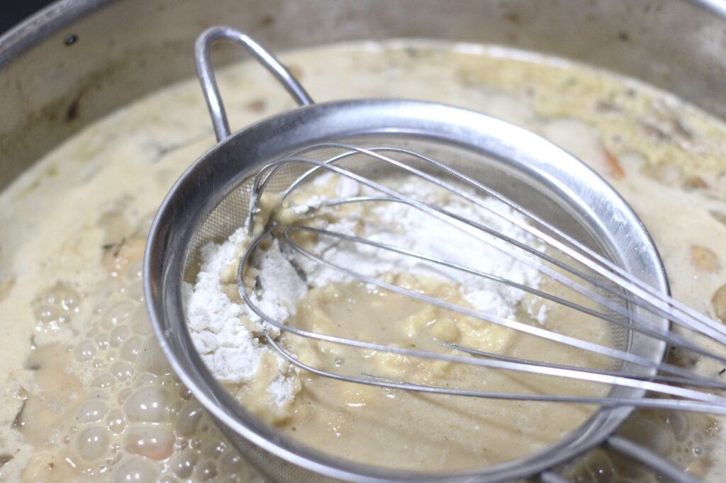 flour being whisked into sauce using a mesh strainer to prevent lumps and thicken the sauce