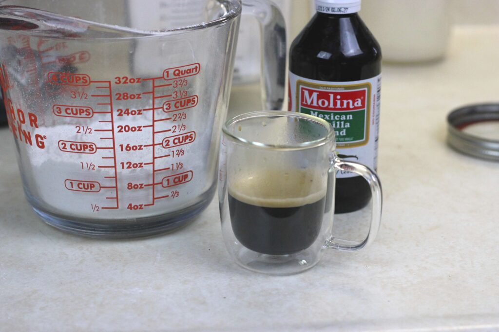 glass measuring cup with powder sugar, bottle of vanilla extract, small glass mug of espresso 